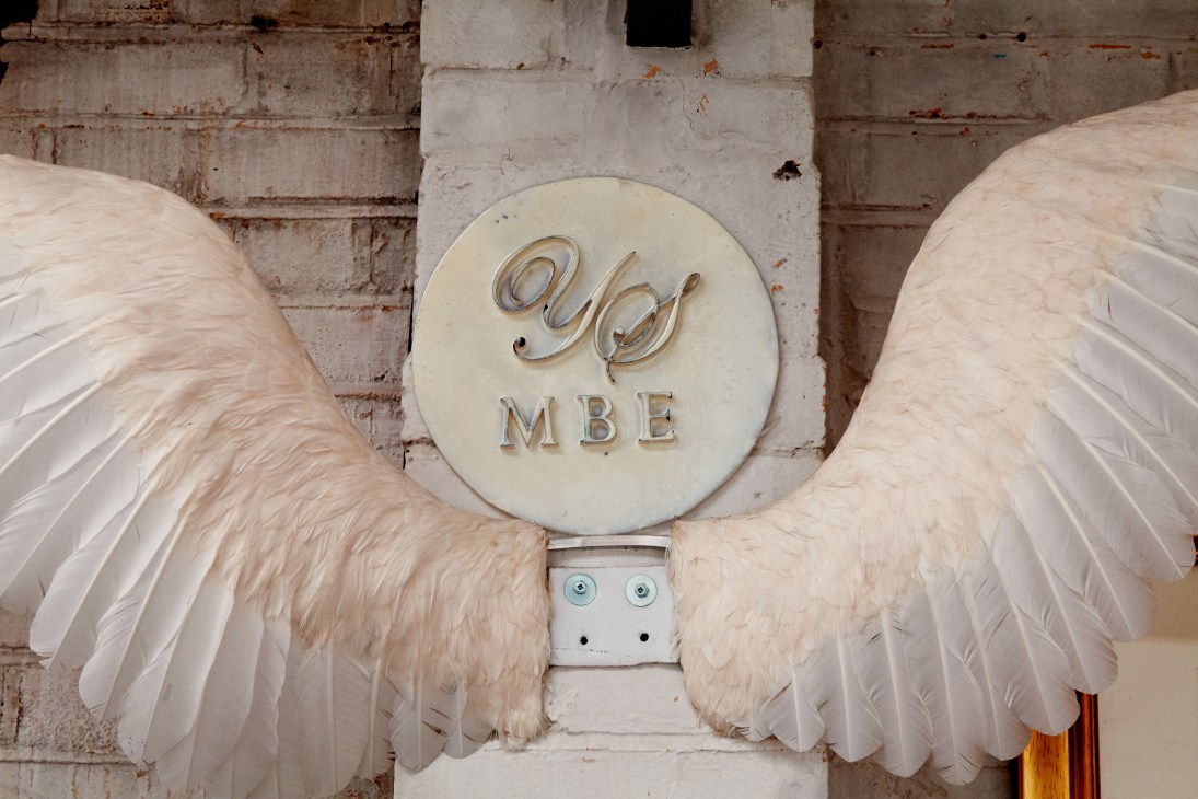 White feathered wings flanking a cream-colored circular plaque engraved with 'YS MBE' mounted on a brick wall.
