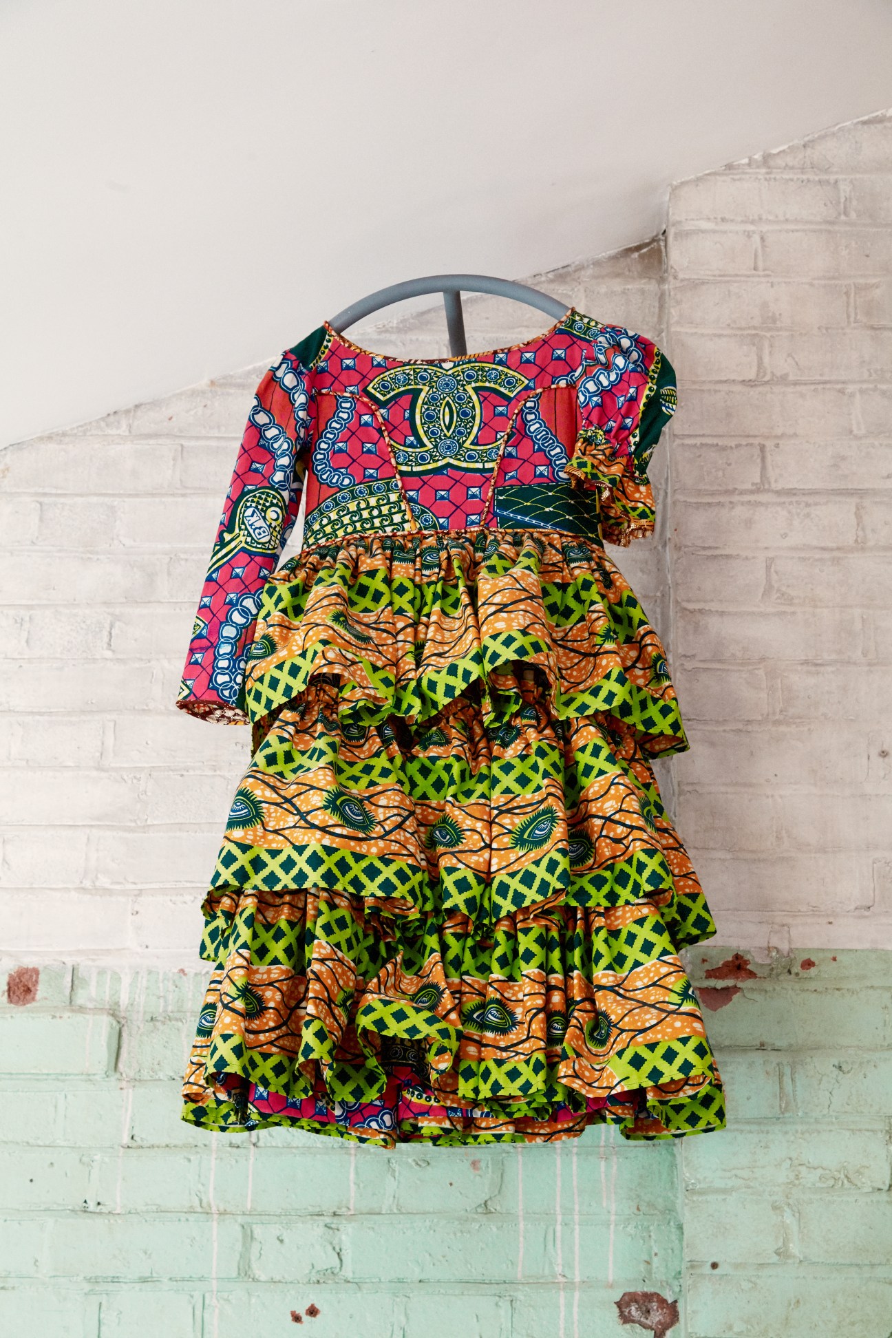 Colorful tiered dress made from patchwork batik fabric in red, green, orange and yellow patterns hanging on a metal hanger against a white brick wall.