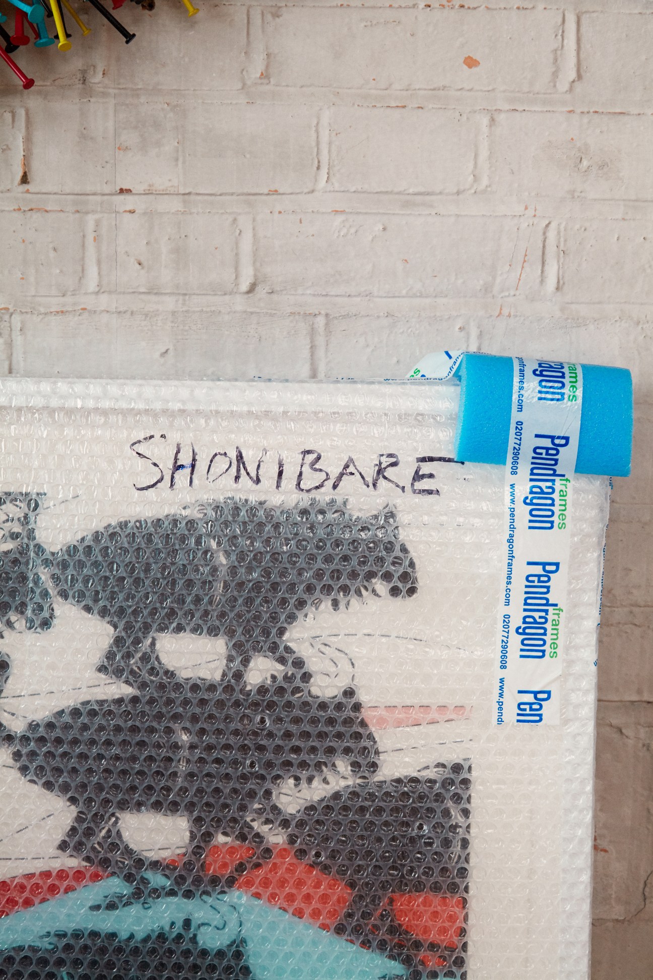 A wrapped print labeled 'SHONIBARE' with bubble wrap packaging and blue shipping label against white brick wall.
