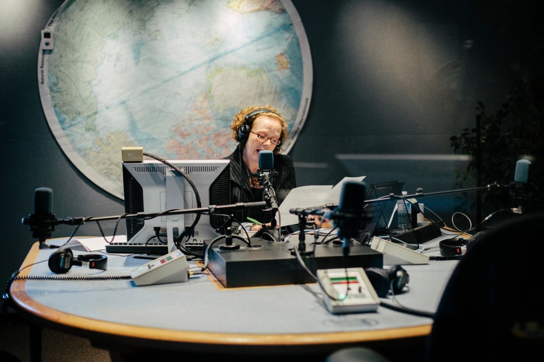 CBC North reporter Sonja Koenig speaks into a microphone at a radio desk with a large globe map behind her.