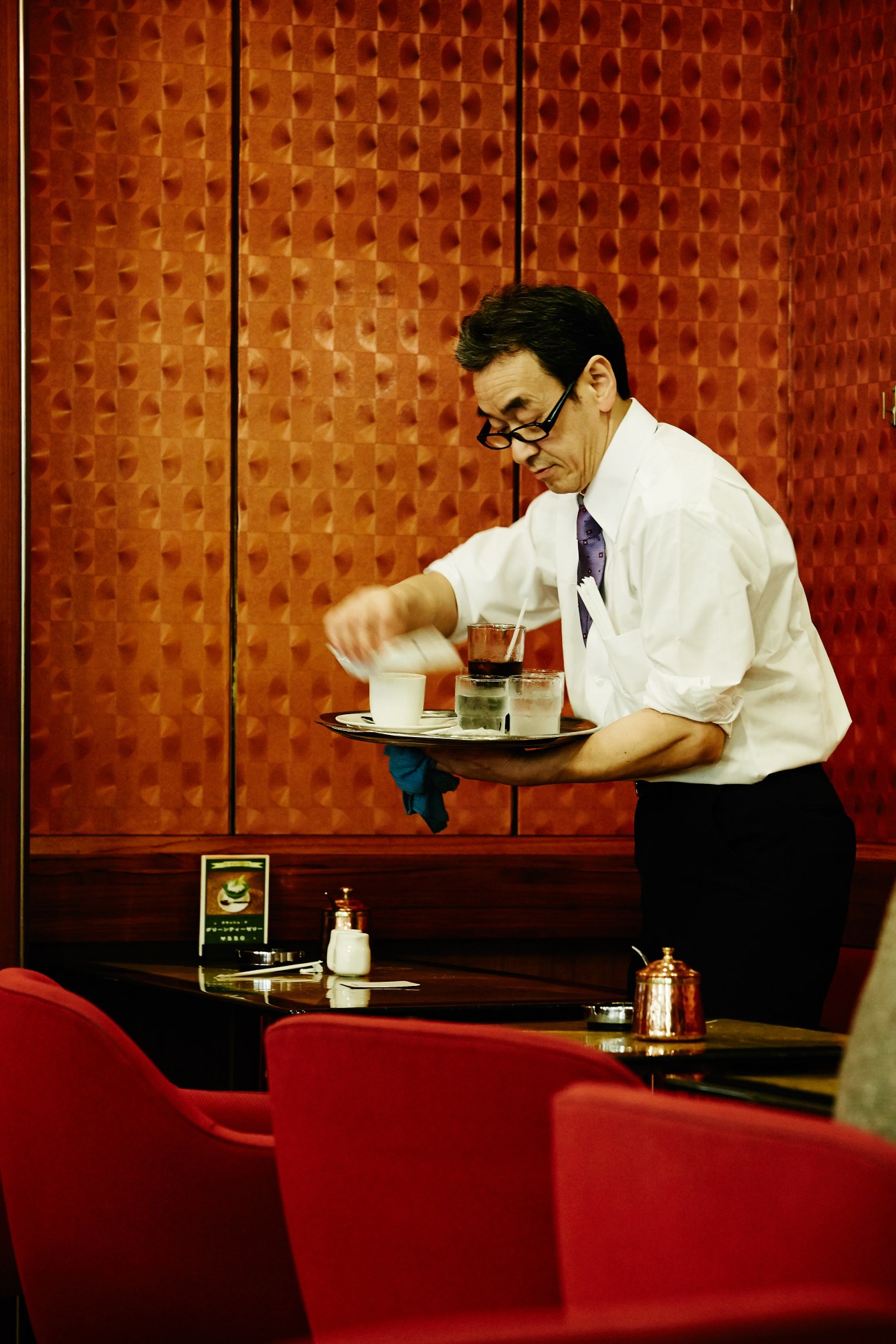 Waiter in white shirt and glasses serves coffee at a retro Tokyo café with red upholstered seating.