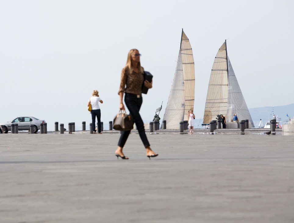 People strolling on a waterfront plaza with sailboats and mountains visible in the background.