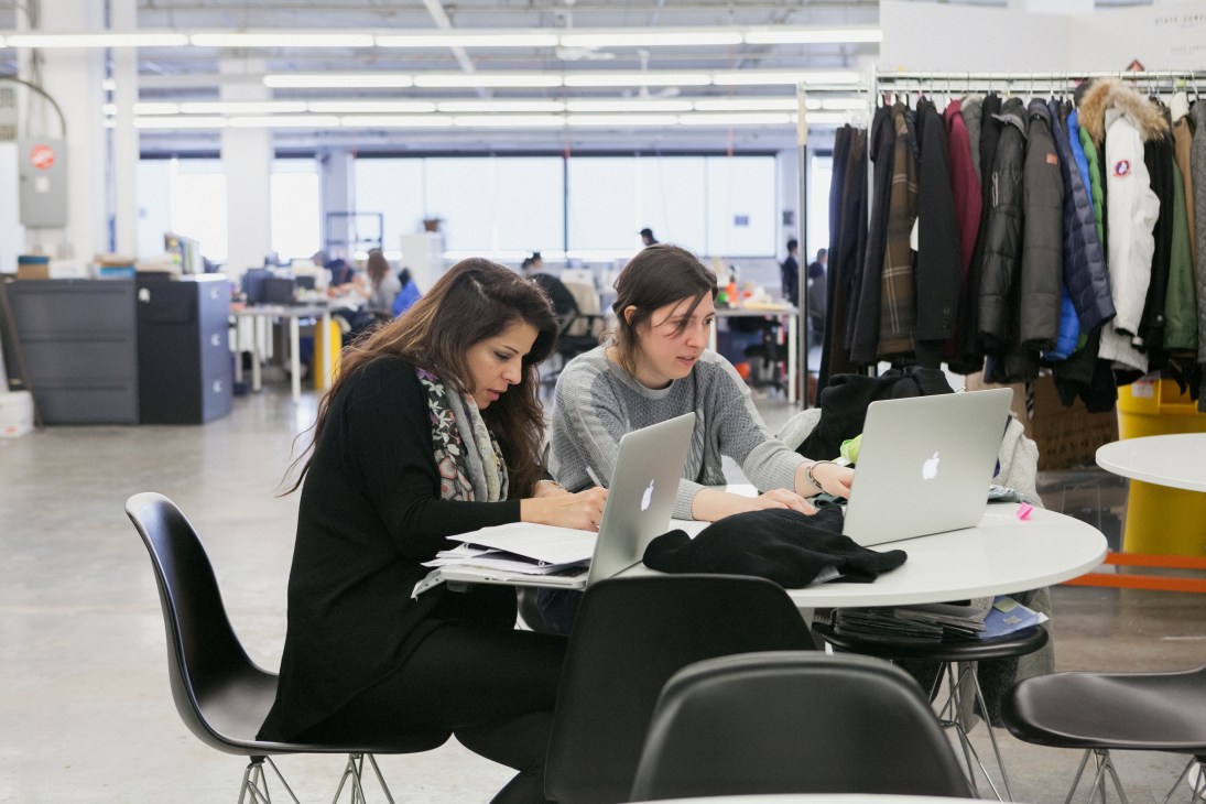 Two women working on laptops in a bright, modern fashion manufacturing office with clothing racks visible in the background.