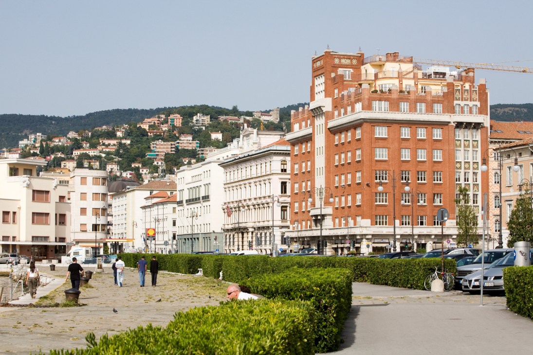 Historic waterfront buildings in Trieste with hills in background and pedestrians in plaza.