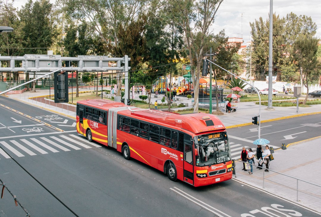 Red Metrobús rapid-transit bus on a street with green space, pedestrian walkways and cycle lanes.