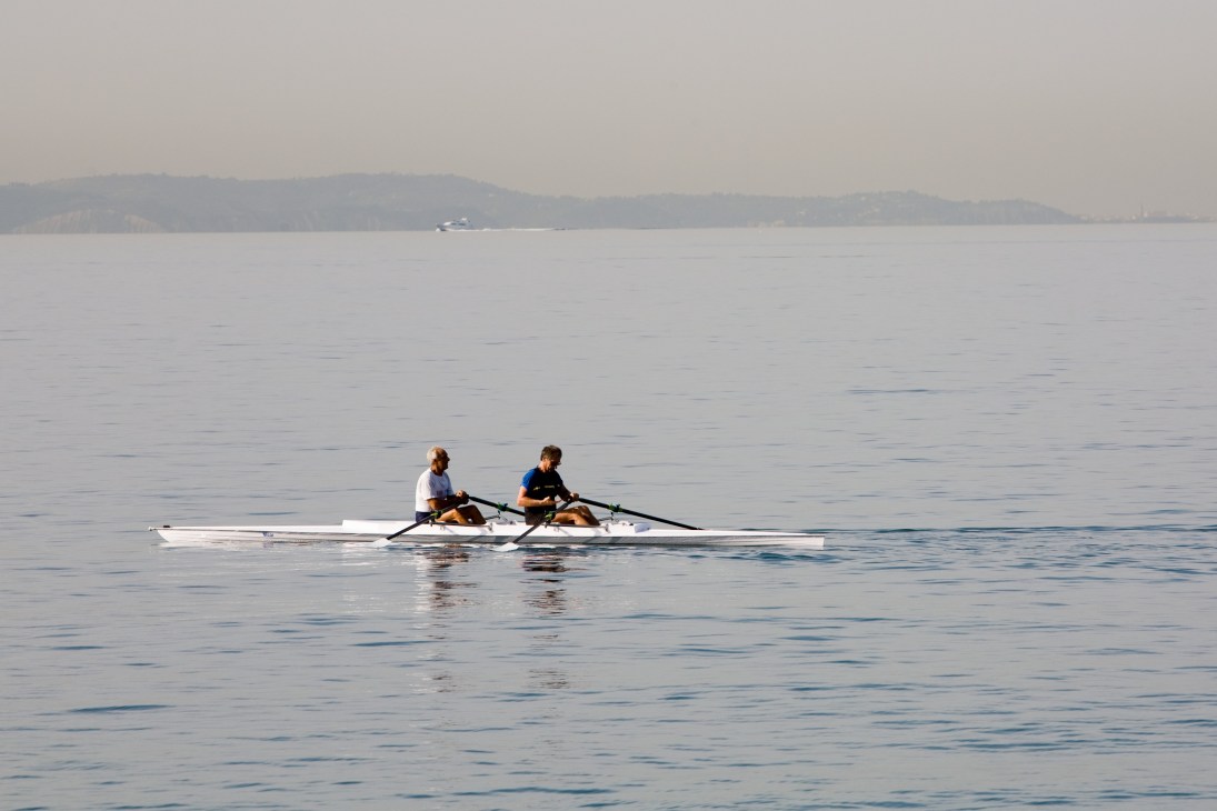 Two people rowing a small boat on calm Adriatic waters with forested hills in the background.