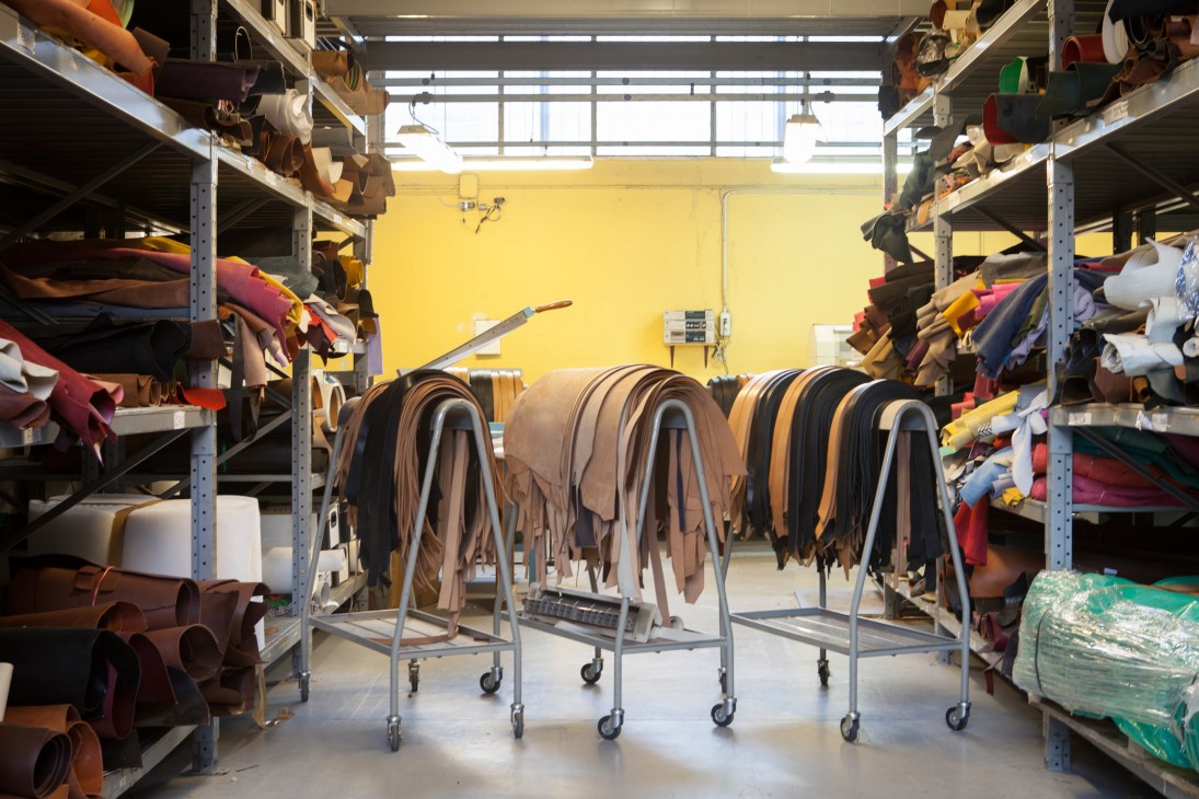 Leather hides hanging on rolling racks in Adriano Meneghetti's workshop with metal shelving and yellow walls.