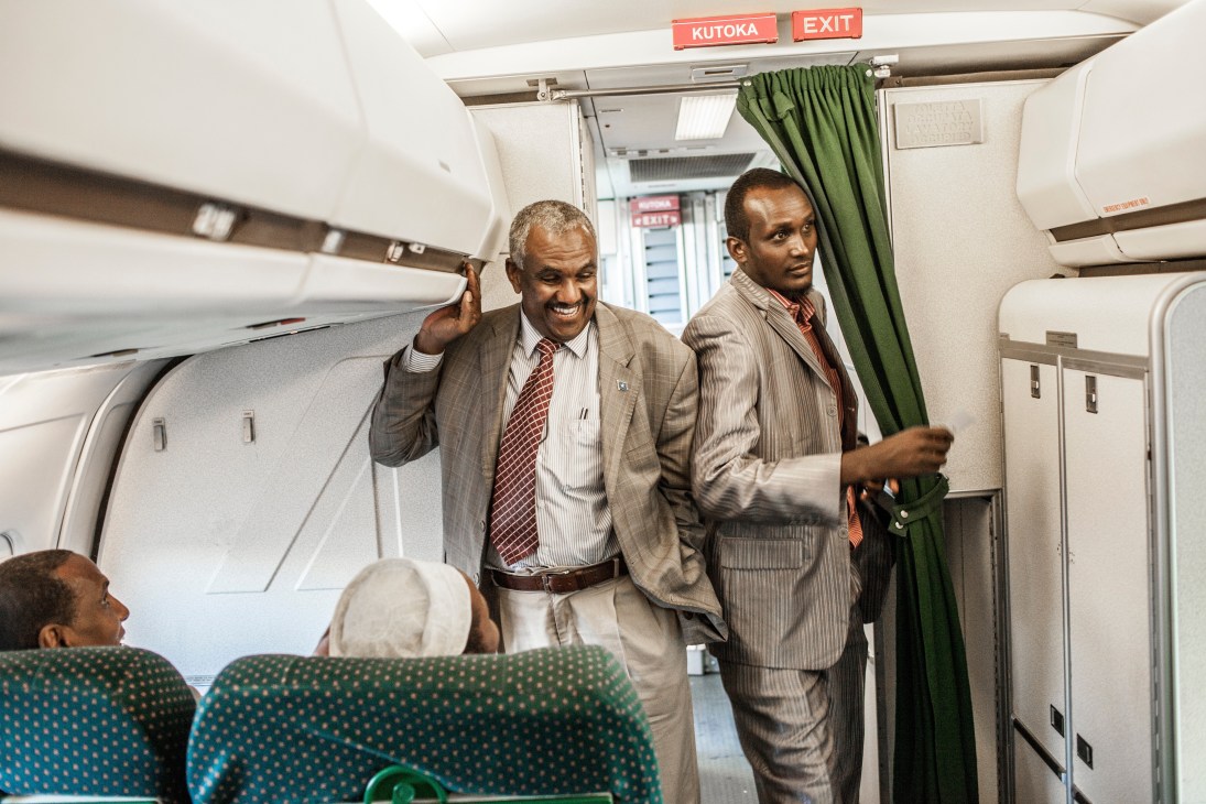 Two men in business suits walking through an aircraft cabin during boarding, with exit sign visible above.