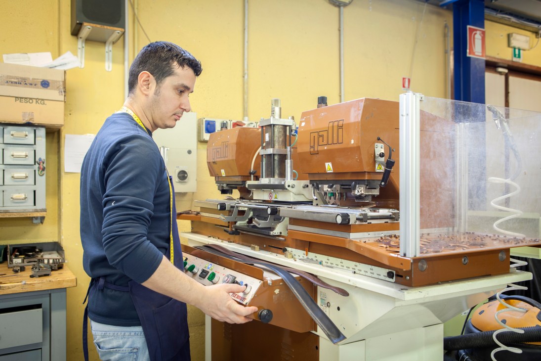 Craftsman operating an industrial belt hole-punch machine in a yellow-walled workshop.