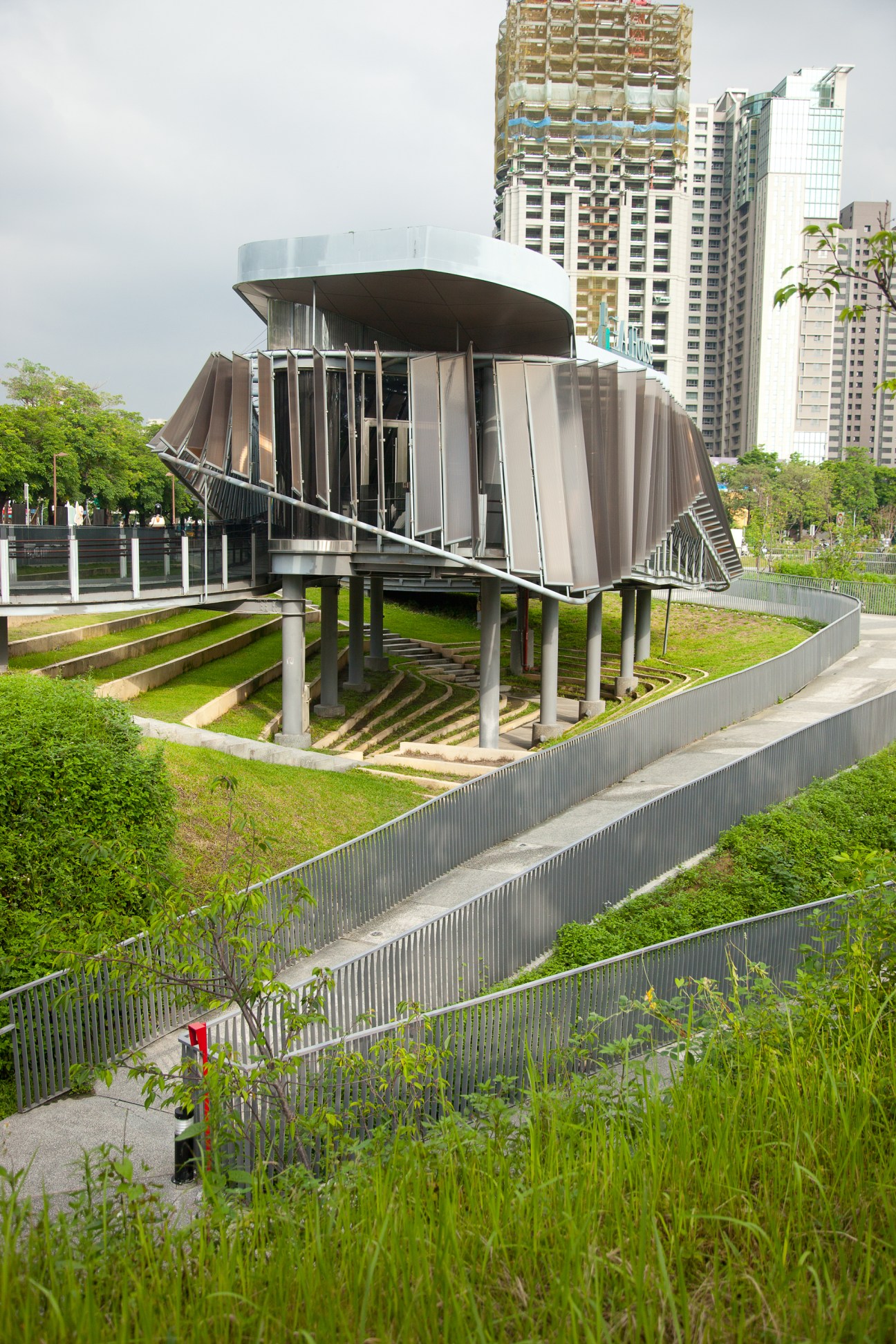 Modern architectural structure with curved metal panels elevated on stilts in a green park setting surrounded by tall buildings.