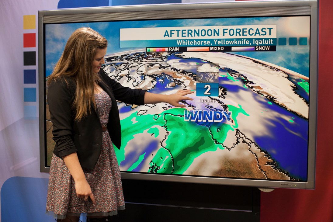 Weather presenter pointing at afternoon forecast map showing windy conditions for Yellowknife on large studio screen.