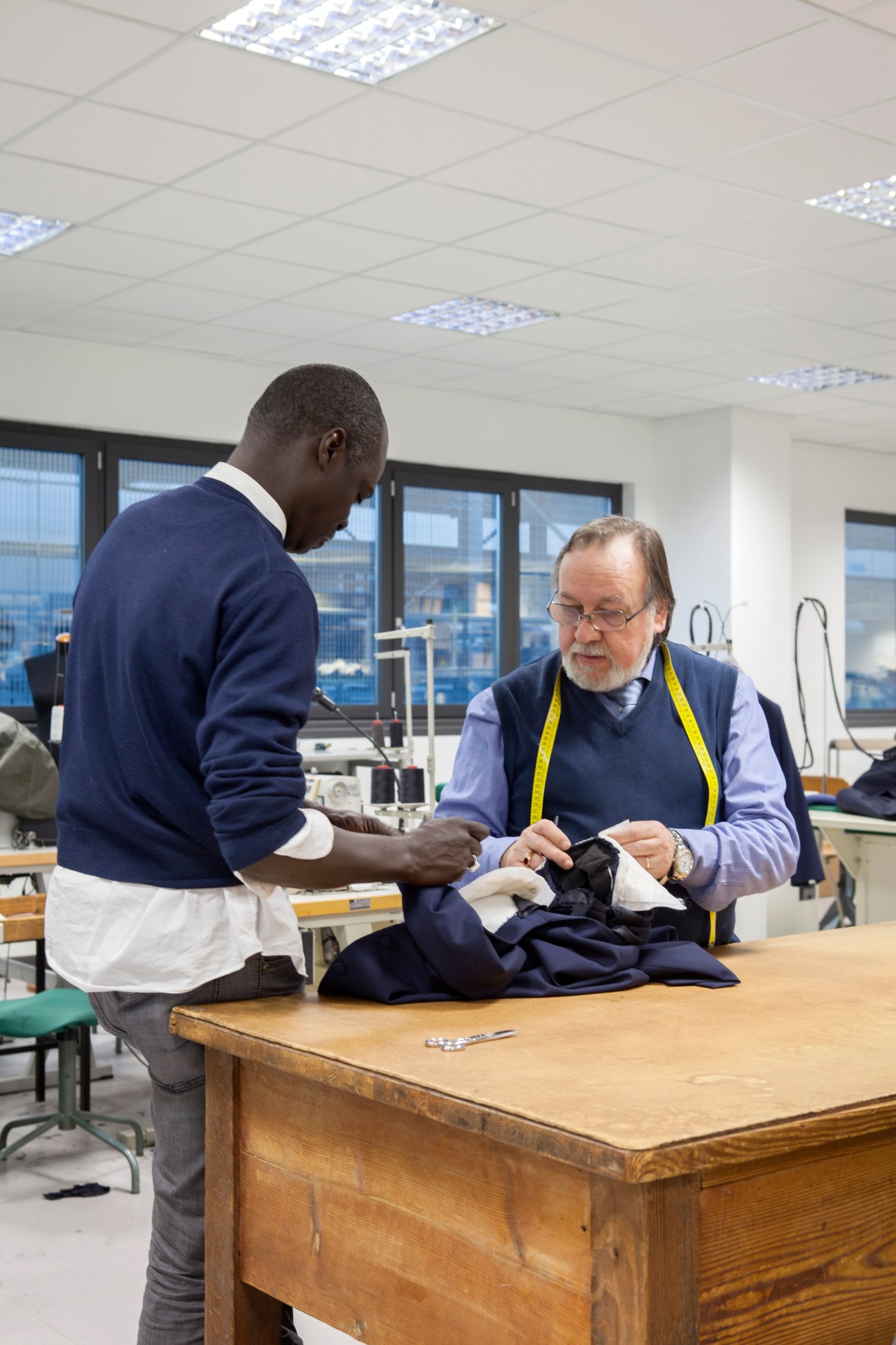 Two men examining dark fabric at a wooden table in a bright workshop with floor-to-ceiling windows overlooking Milan.