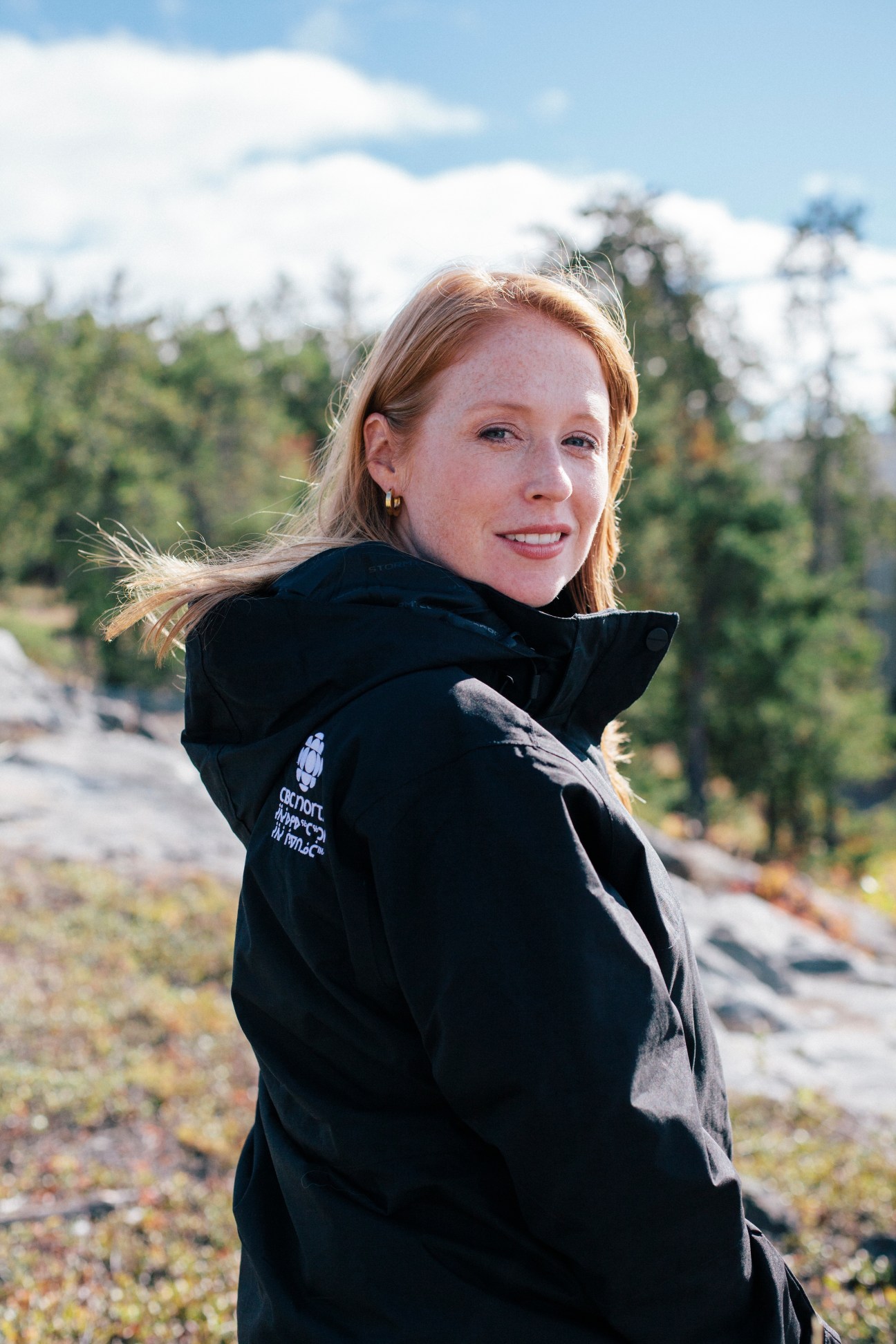 CBC North reporter Kate Kyle smiling at camera outdoors with boreal forest and rocky terrain behind her.