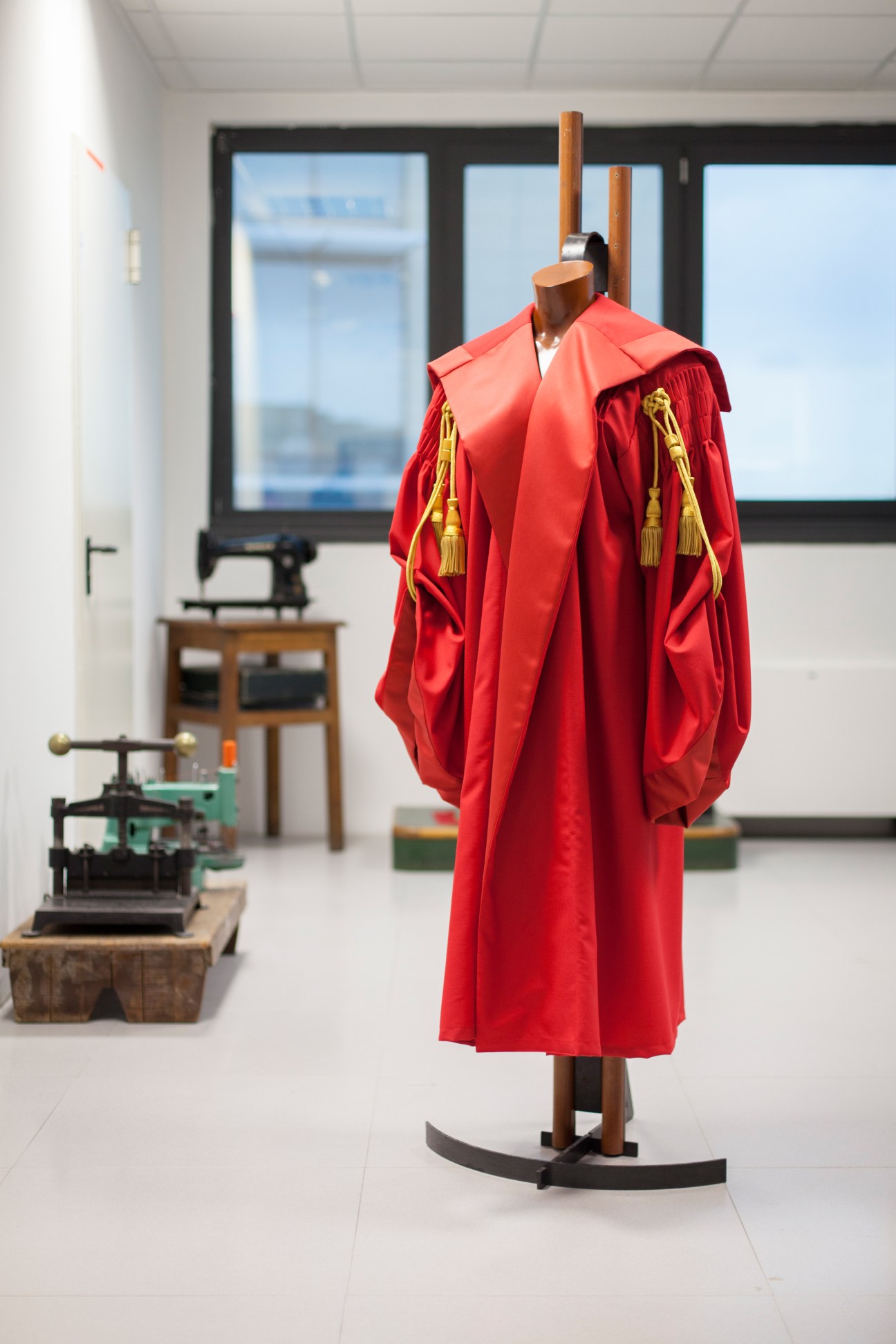 Red judge's robes with gold tassels displayed on a mannequin in a bright showroom with vintage tailoring equipment visible.