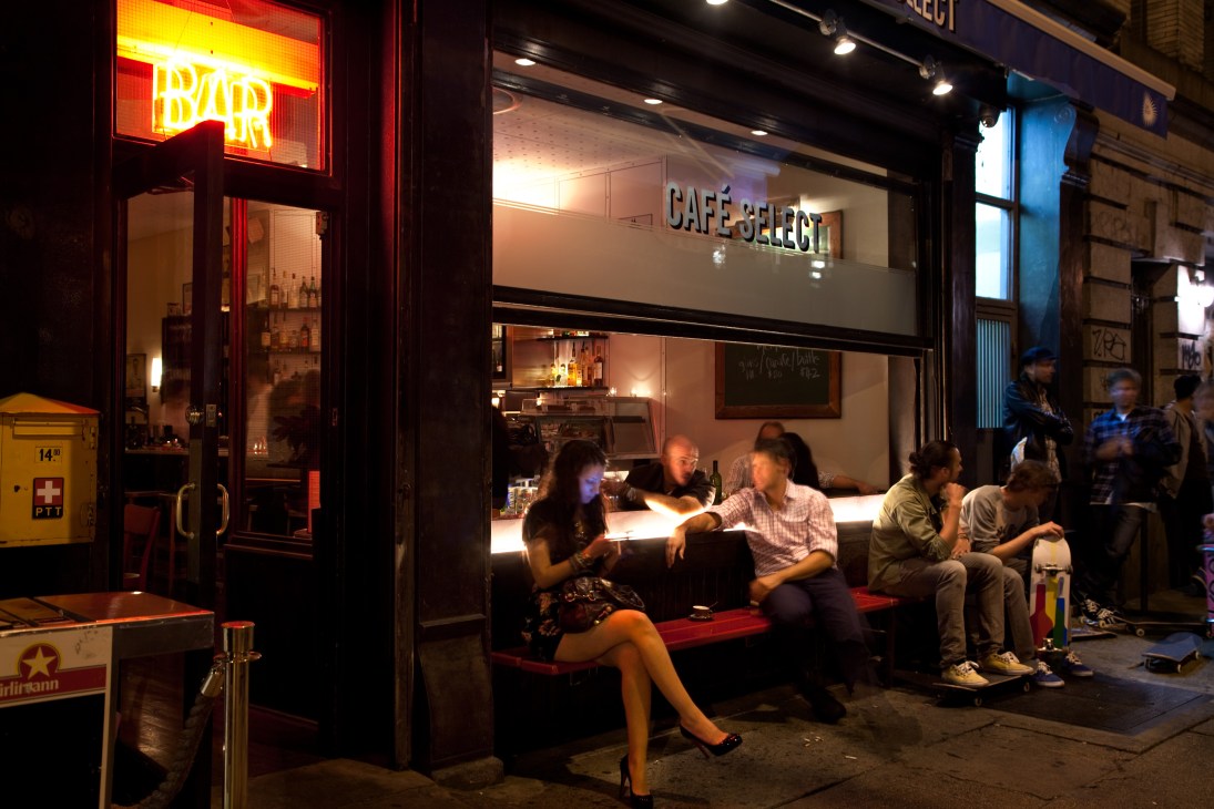 Café Select storefront in Manhattan's NoLita at night with patrons seated outside on a red bench.