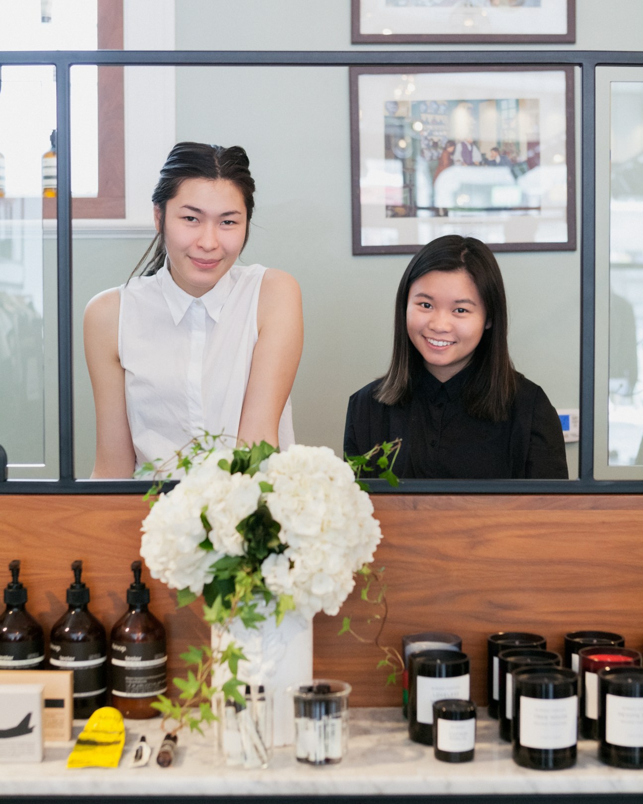 Two women stand behind a wooden counter at Want Apothecary, with white flowers and dark glass bottles displayed in front.