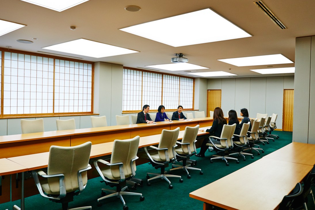 A modern meeting room with wooden tables, beige chairs, and a panel of three officials seated at the head of the table.