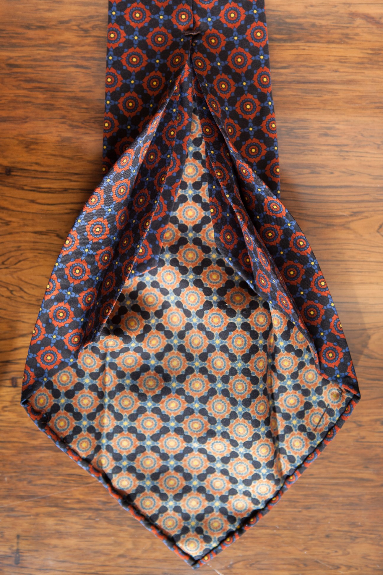 A seven-fold silk tie by Petronius displayed flat on wood, showing an orange and navy geometric pattern.