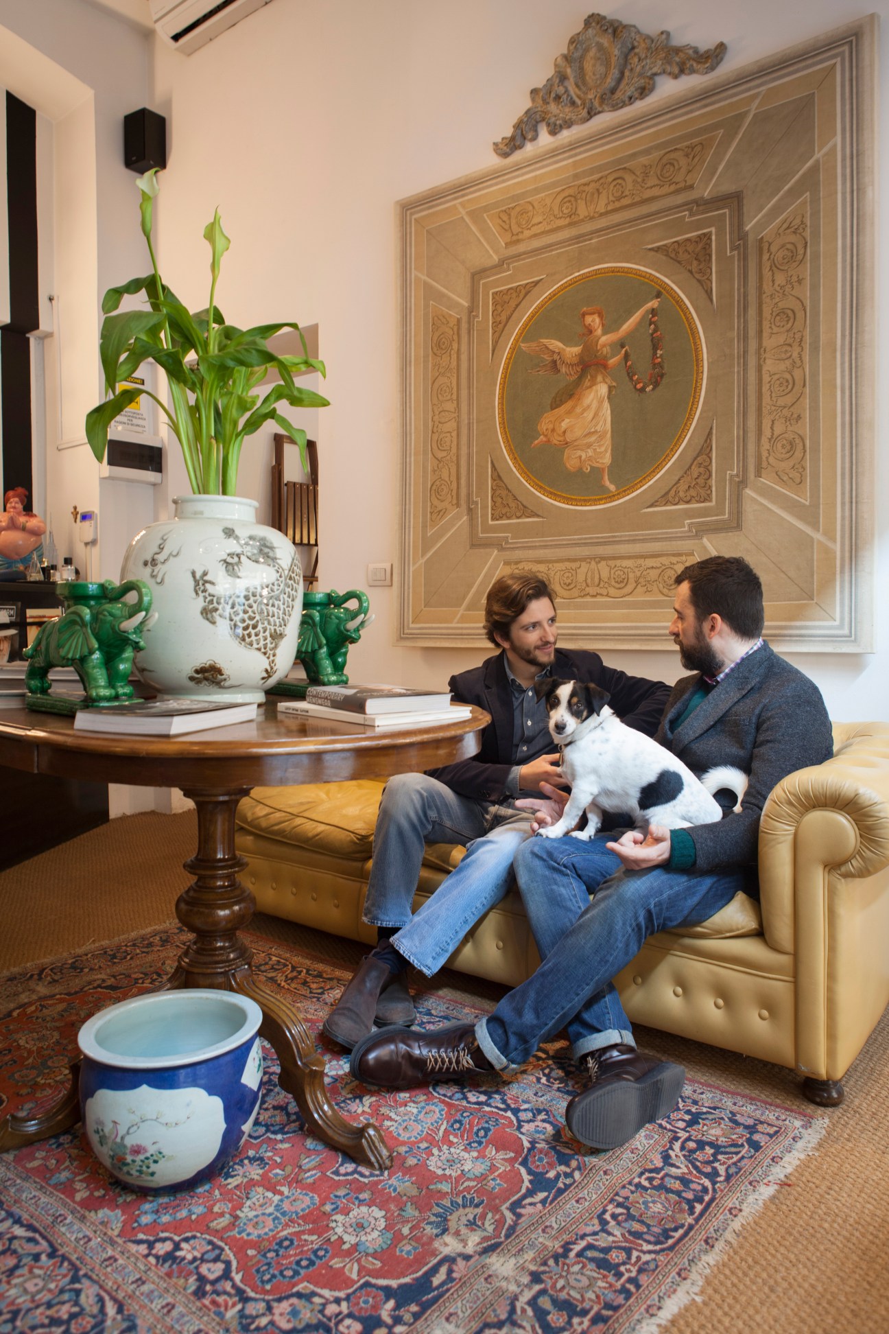 Two men sit on a cream leather sofa holding a black and white dog in an elegant interior with classical artwork and decorative objects.