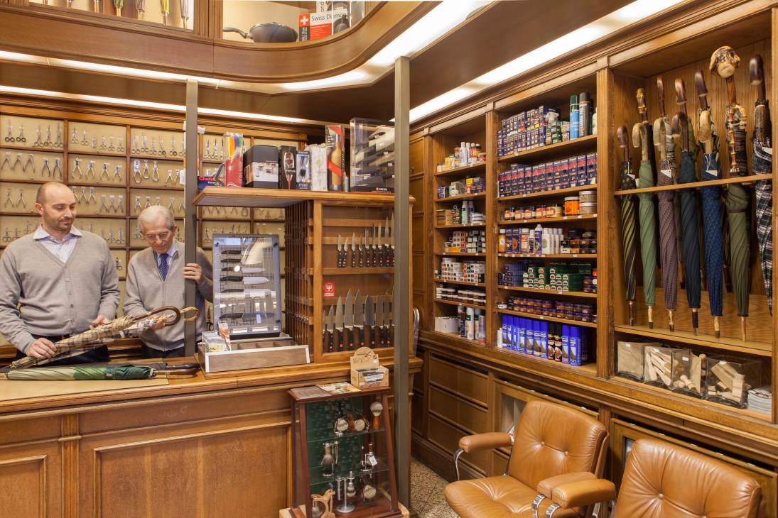 Two men stand in a traditional wooden shop interior with shelves displaying umbrellas, tools, and grooming products.