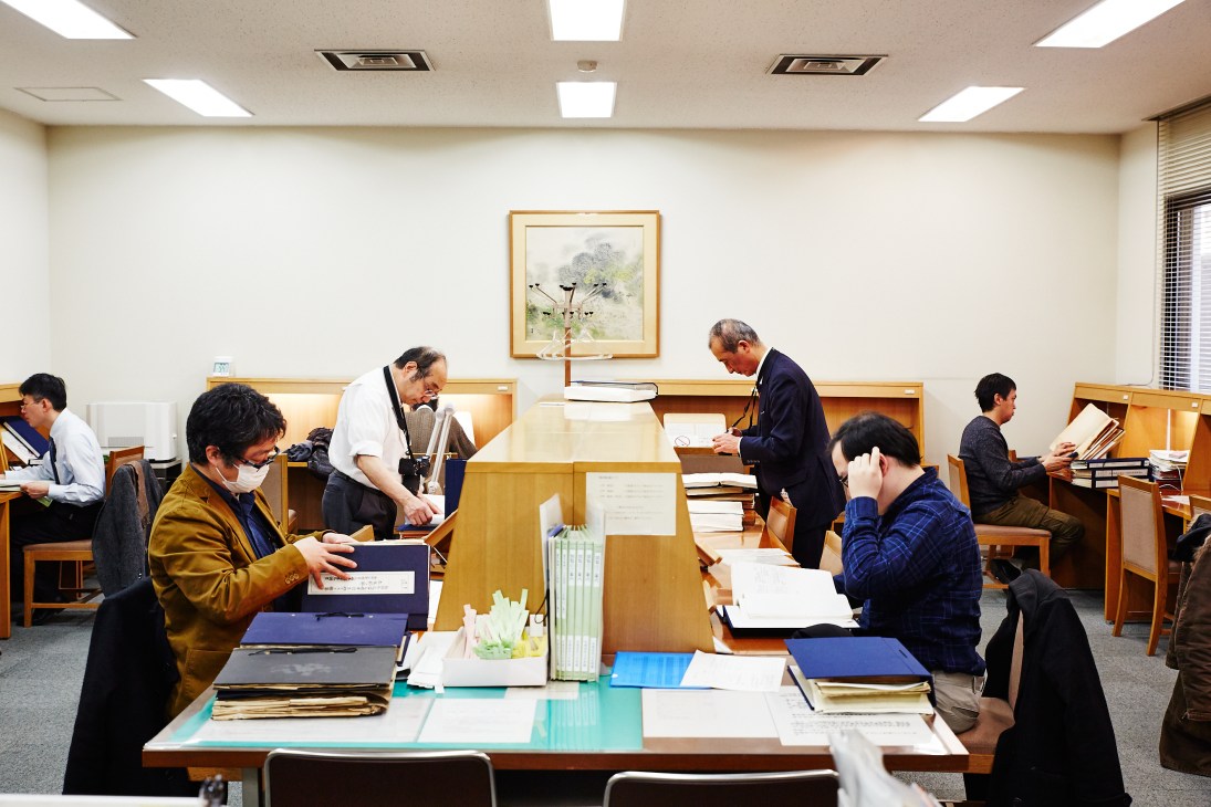 Multiple people working at desks and shelves in the ministry's Diplomatic Archive reading room with wooden furniture and artwork on walls.