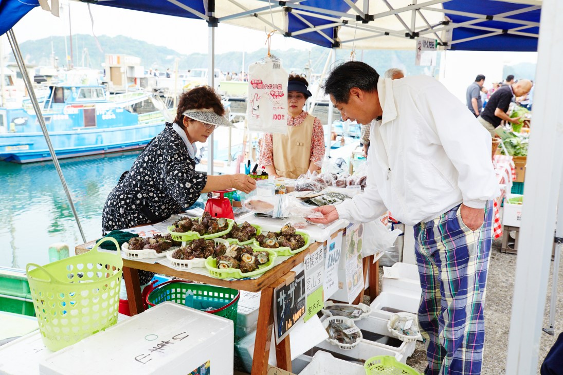 Vendors and customers at Hayama's weekly farmers' market with fresh shellfish displayed under blue umbrellas by the fishing port.