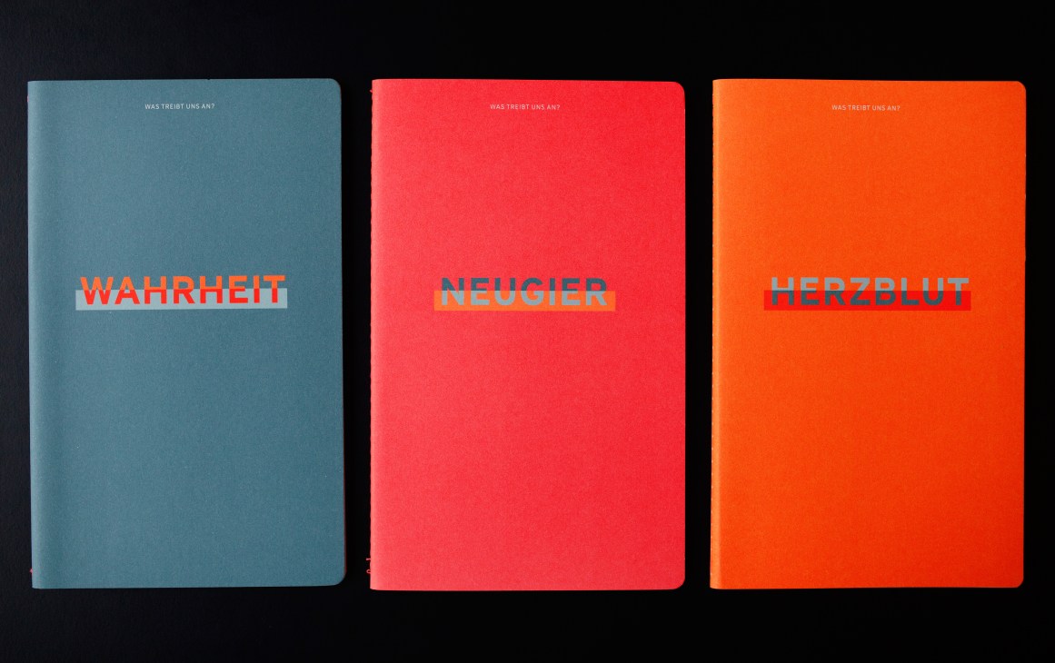 Three colorful German-language notebooks in blue, red, and orange with contrasting text reading 'Wahrheit,' 'Neugier,' and 'Herzblut.'