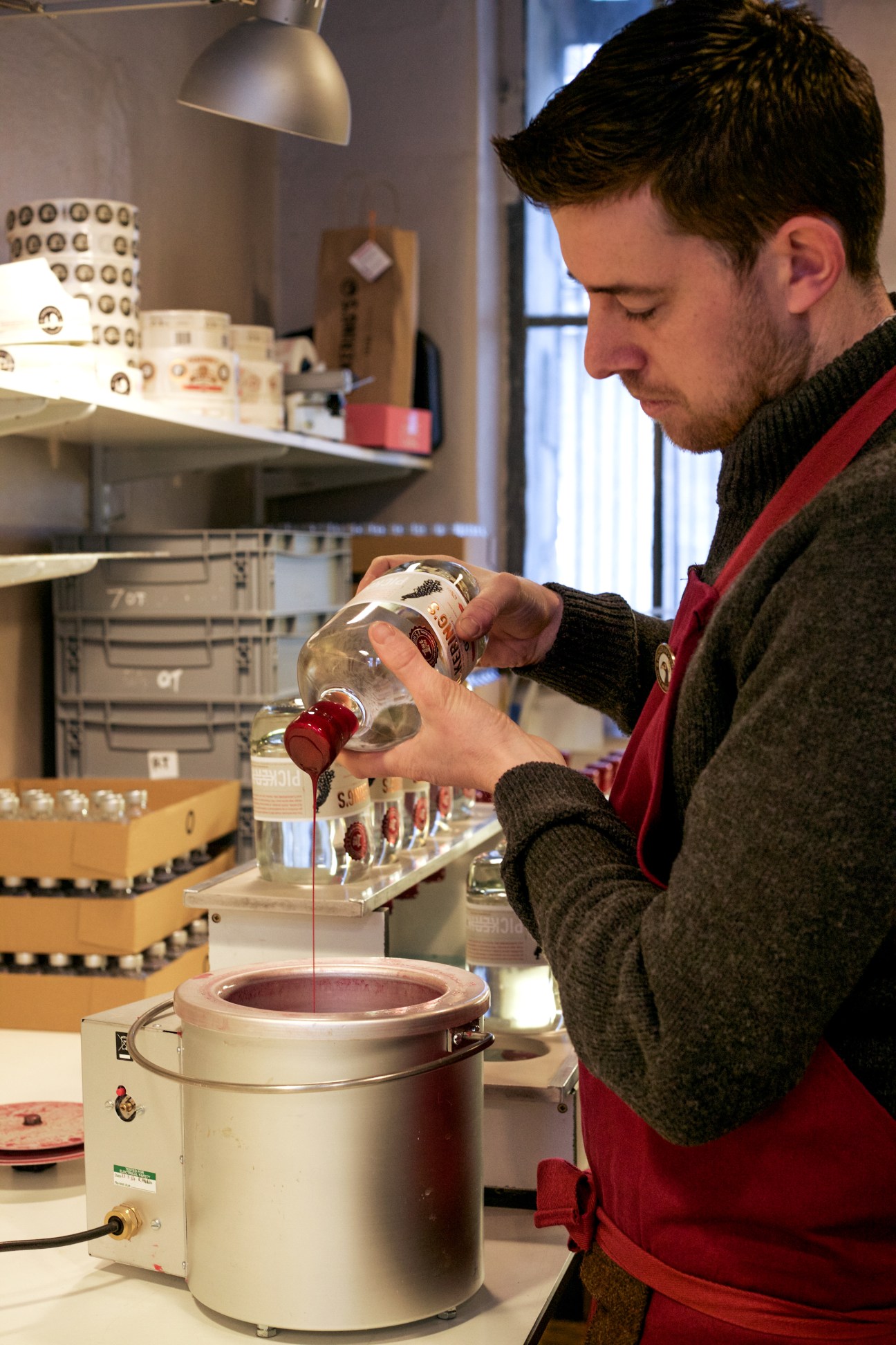 A man in a red and black sweater pours liquid wax from a bottle into a container during gin production.