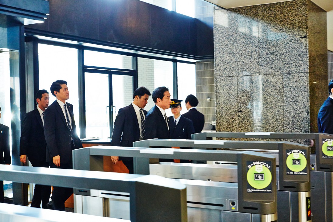Foreign minister Fumio Kishida leads Japanese officials through a modern building entrance with security gates.