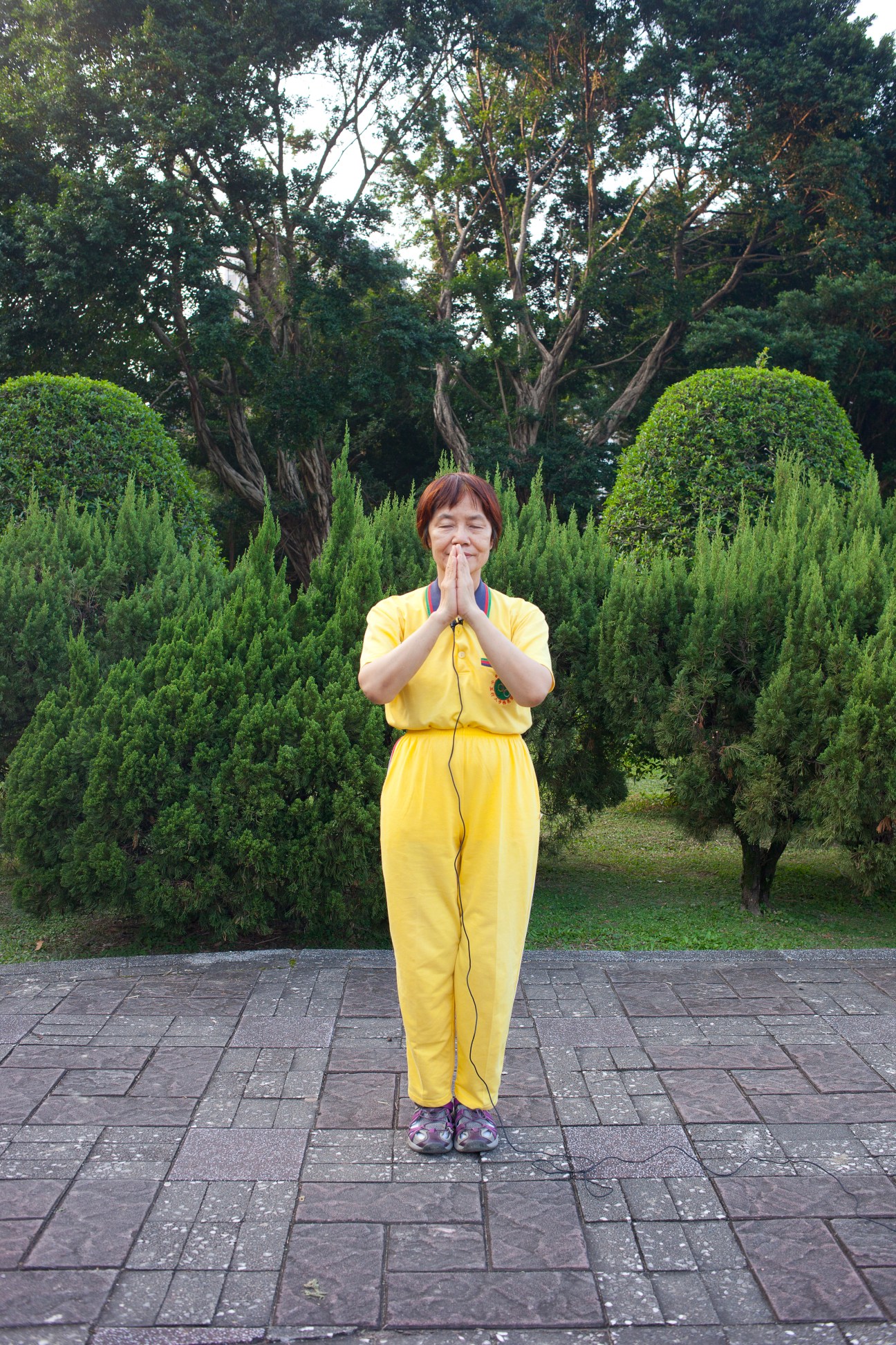 Woman in yellow outfit meditating with hands pressed together in a garden with tall trees.