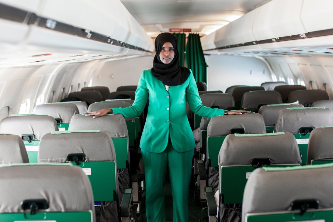 African Express stewardess Hodho Abshir in green uniform standing in aircraft cabin aisle.