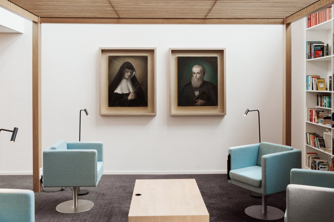 Two framed portraits hanging in a library with shelving, depicting the founders of Regina Coeli language school.