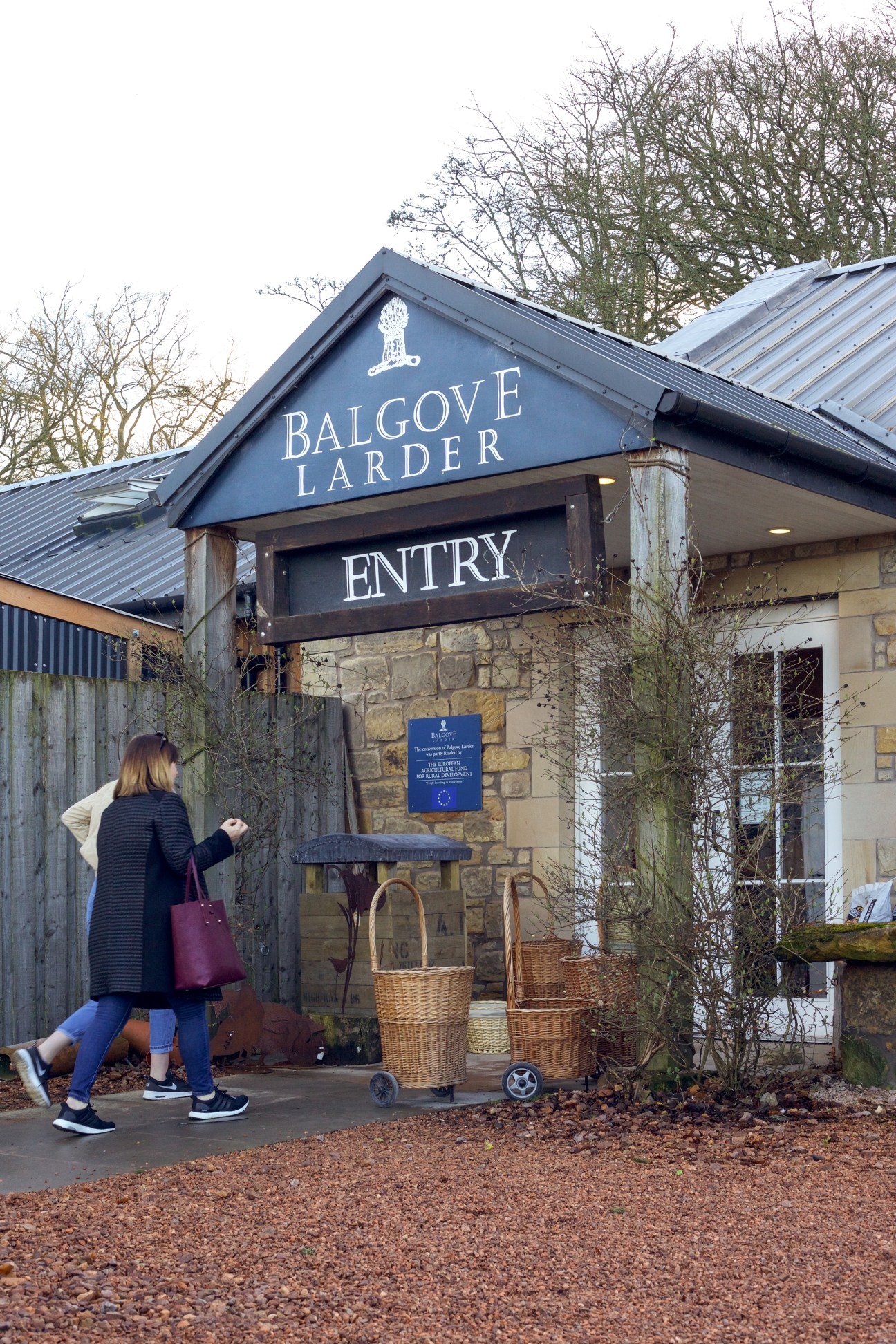 A woman approaching the entrance to Balgove Larder, a farm shop with a dark blue gable sign and wicker baskets outside.