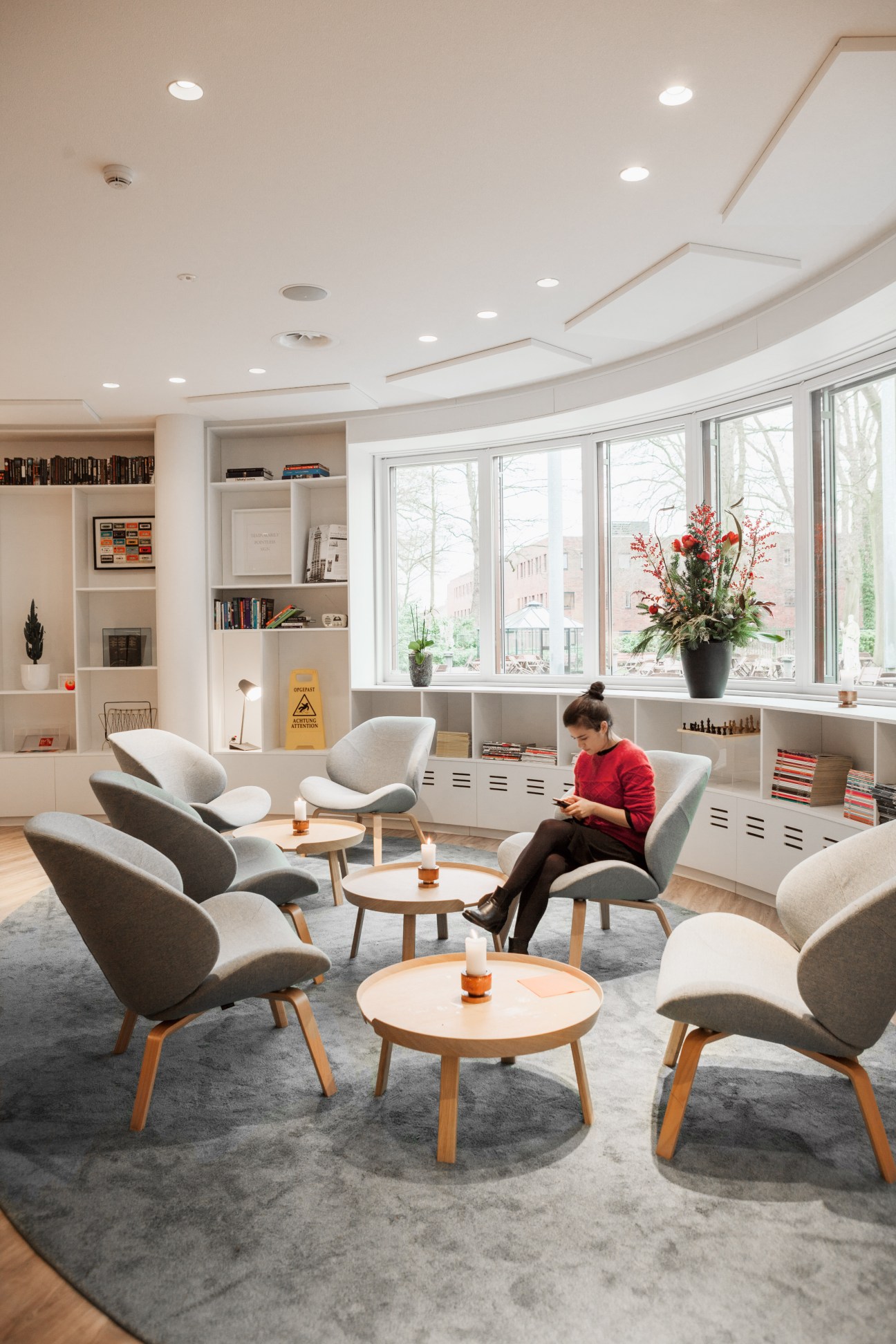 Modern lounge area with grey armchairs, wooden tables, and large windows overlooking a city street.