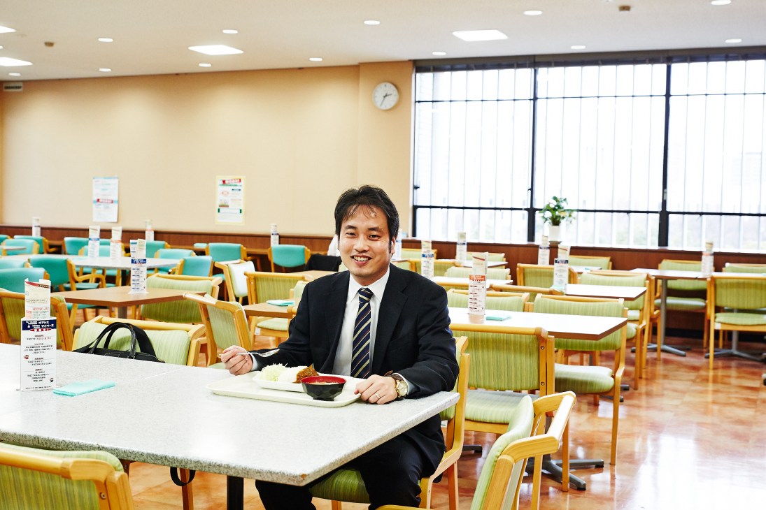 Press officer Toshiya Takarada sitting at a table in a bright cafeteria with green chairs.