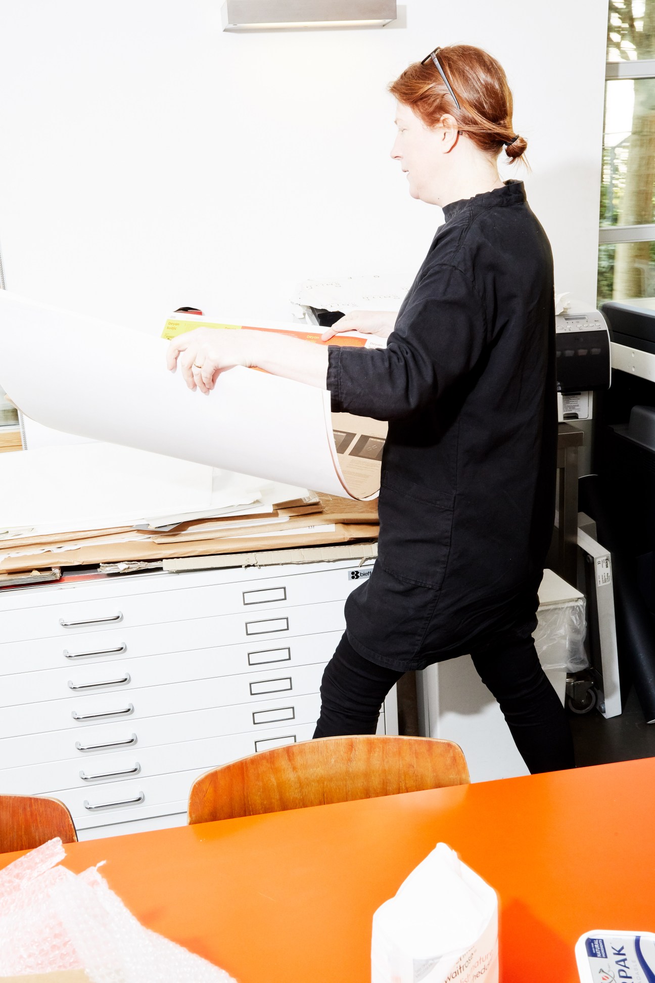 Woman with reddish hair wearing black jacket holding rolled papers in design studio with filing cabinets.