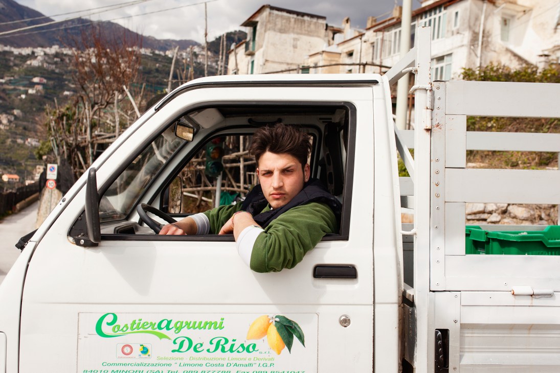 A truck driver sits in the cab of a white delivery van marked with lemon branding on the Amalfi Coast.