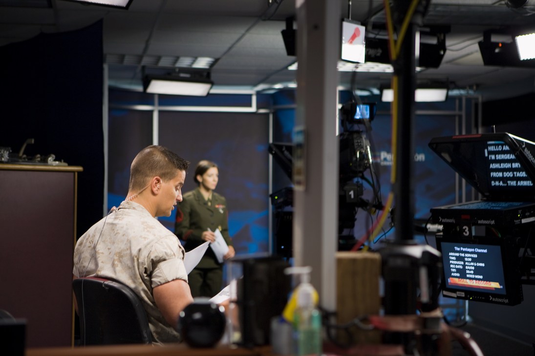 A service member in a light-colored t-shirt holds papers while preparing on a television studio set, with military personnel visible in the background.