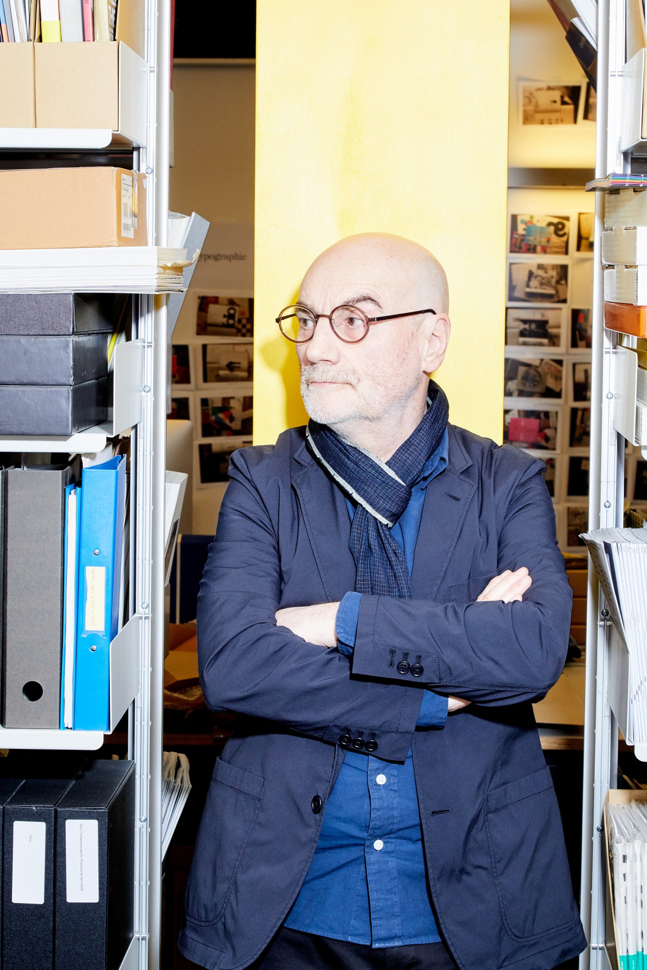 Adrian Shaughnessy, Unit Editions co-founder, standing with arms crossed in a bright yellow office space lined with shelves.