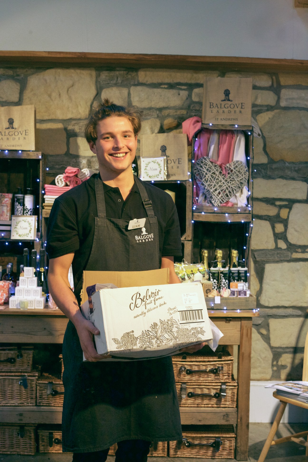 Staff member Liam smiling while holding a Balgove Larder box in the café, with stone walls and retail displays behind him.