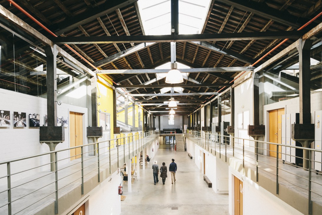 Interior of BAU design school showing industrial warehouse conversion with exposed beams, yellow doors, and people walking through open space.