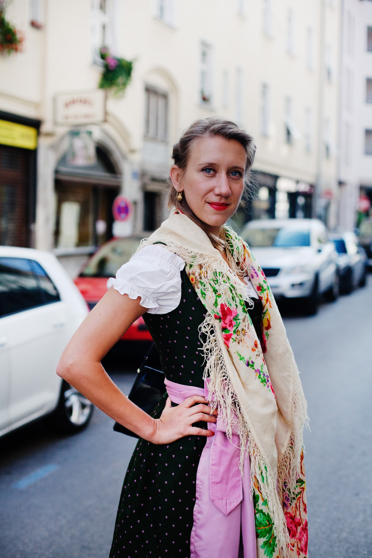 Woman wearing traditional Bavarian dirndl with black dress, white puffed sleeves, pink bow, and floral shawl on a Munich street.