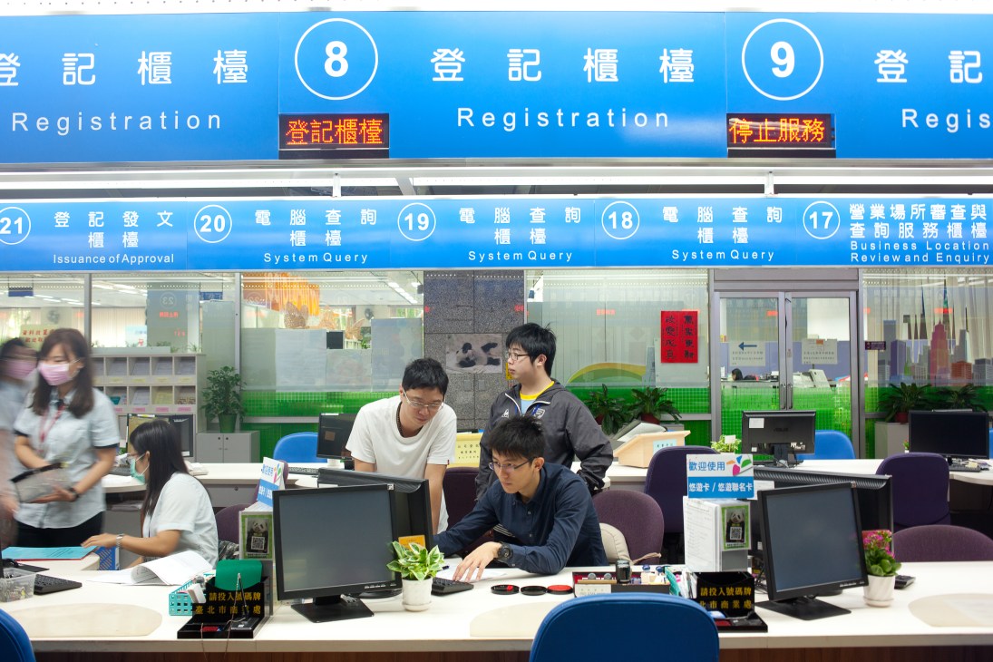 Staff work at computer stations inside a modern Taipei city hall office with blue signage and Chinese characters on the wall.