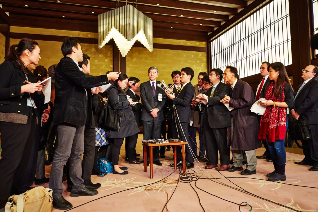 Media scrum of journalists and photographers surrounding officials at a microphone in an elegant room with gold walls.