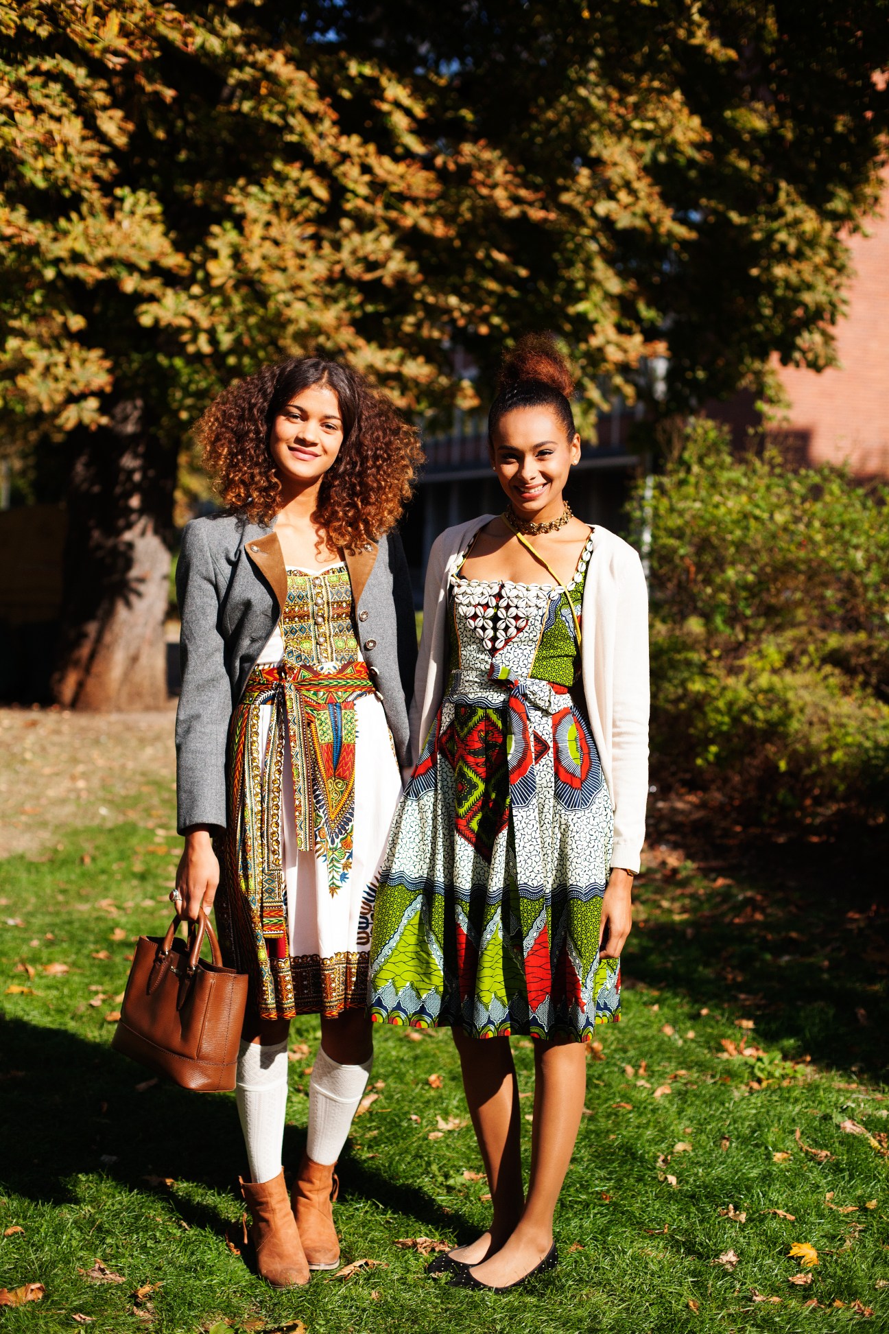 Two young women wearing traditional Bavarian dirndl dresses with embroidered details and cardigans standing outdoors on grass.