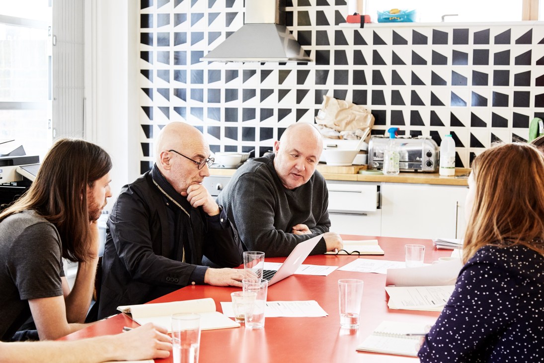 Four people seated at a red table in a modern office with geometric patterned wall shelving during an editorial meeting.