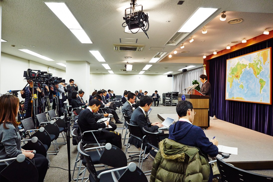 Press secretary Yasuhisa Kawamura stands at a podium addressing journalists and camera crews in a conference room.