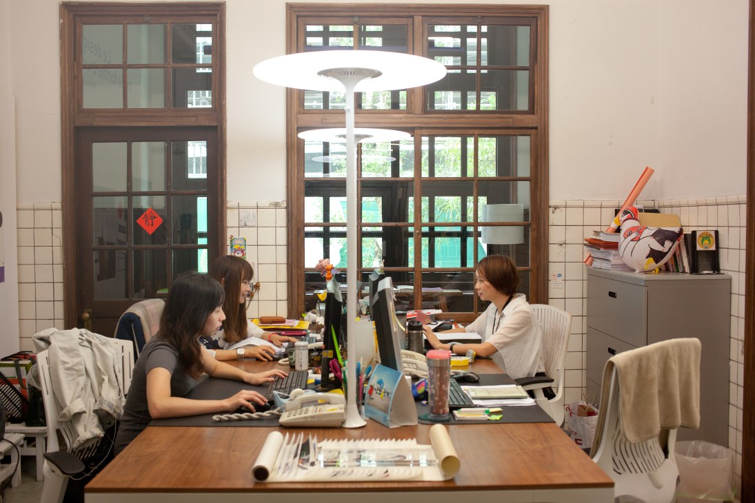 Two women work at desks in a bright office with large wooden-framed windows and creative materials scattered about.
