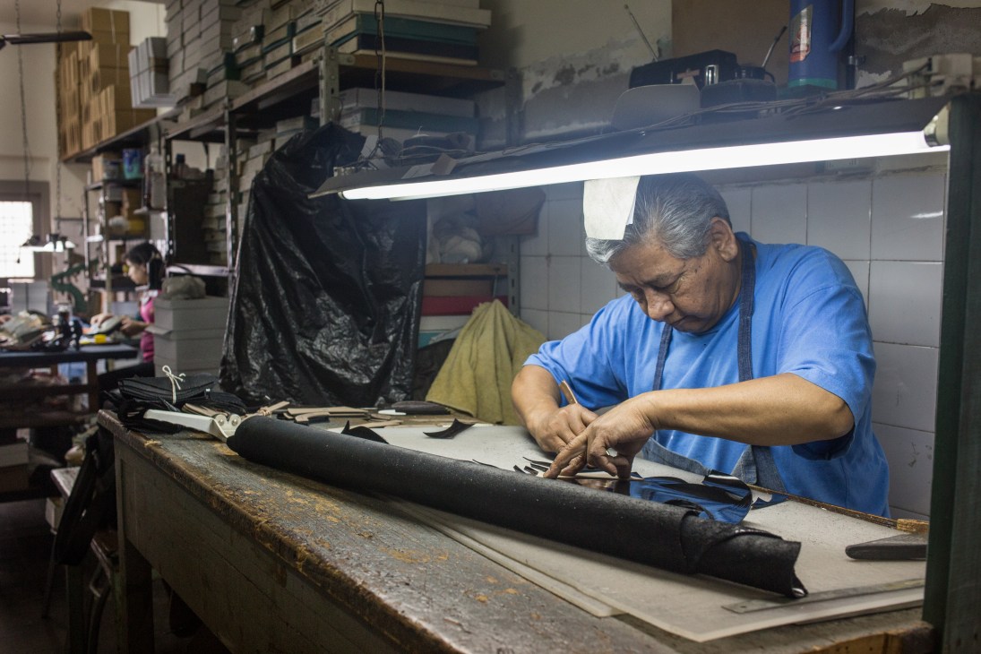 Craftsperson in blue shirt hand-stitching black leather tango shoe at workbench under bright light.