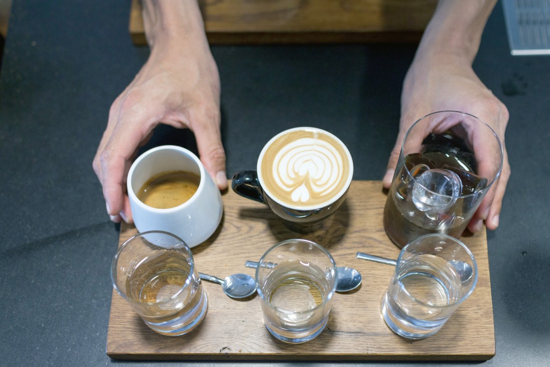 Hands presenting espresso, latte, and water glasses on a wooden tray at Sakona coffee shop.